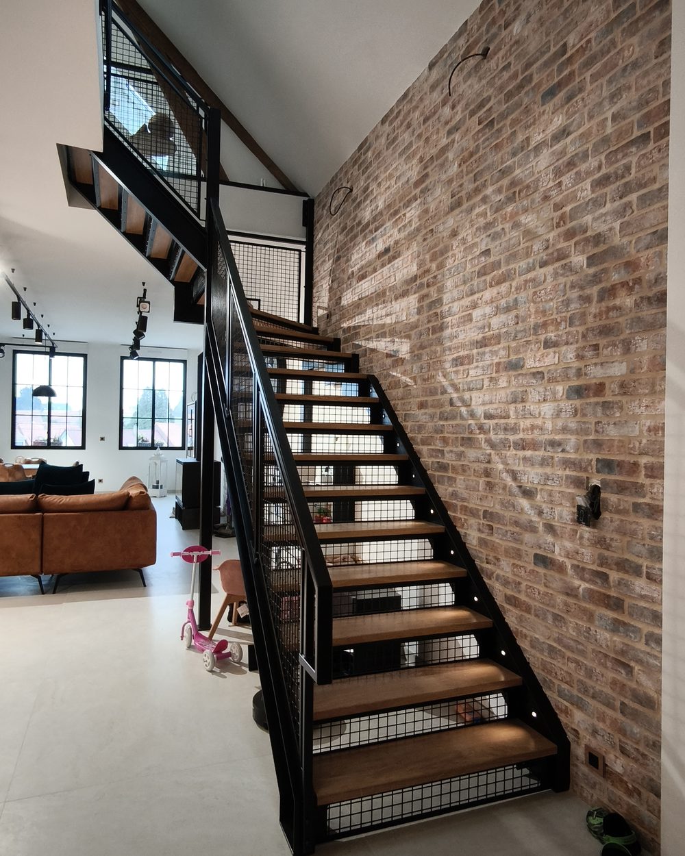 Escalier droit en acier noir et chêne devant un mur de briques, garde-corps en maille d’acier